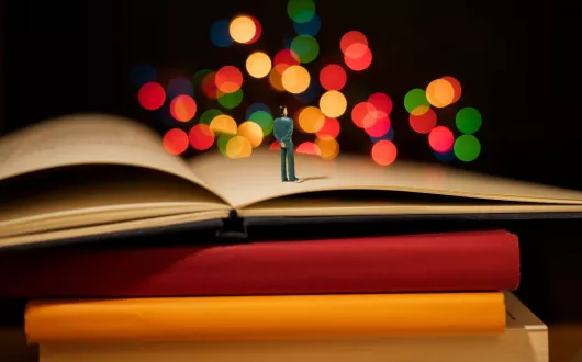 Książki ułożone na stosie. Na otwartej książce leżącej na górze stoi człowiek. 