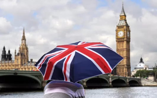 Osoba odwrócona tyłem trzyma parasolkę z motywem flagi Wielkiej Brytanii. W tle widoczne są charakterystyczne londyńskie zabytki – po lewej Pałac Westminsterski, a po prawej wieża zegarowa Big Ben. Przed budynkami przepływa rzeka Tamiza, a na niebie unoszą się jasne chmury.