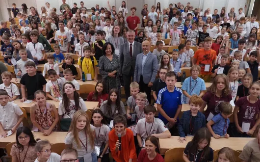 Duża grupa dzieci i kilku dorosłych pozuje do wspólnego zdjęcia w sali wykładowej. Dzieci siedzą i stoją między ławkami, uśmiechają się w stronę aparatu. W pierwszym rzędzie kilka osób dorosłych, w tym kobieta z mikrofonem.