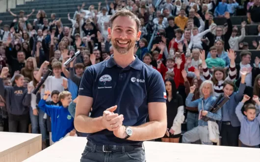 Uśmiechnięty mężczyzna w granatowej koszulce z logo ESA stoi na scenie przed dużą grupą ludzi w sali audytoryjnej. W tle widać wielu młodych uczestników, którzy podnoszą ręce i klaszczą.