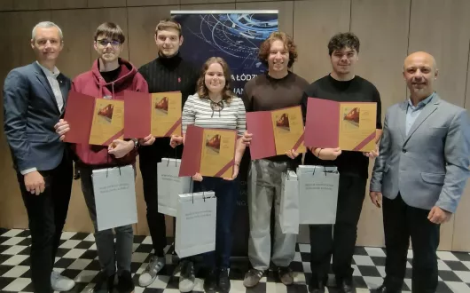 Grupa siedmiu osób pozuje do wspólnego zdjęcia na tle plakatu z logo Politechniki Łódzkiej. Pięć osób w środku, trzy kobiety i dwóch mężczyzn, trzyma dyplomy i torby z napisem „Wydział Mechaniczny Politechniki Łódzkiej”. Po bokach stoją dwaj mężczyźni w garniturach.