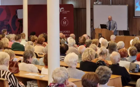 W sali wykładowej trwa wykład dla seniorów. Widzimy wiele osób siedzących na ławkach i słuchających prelegenta, który stoi przy mównicy z mikrofonem i laptopem. Za nim znajduje się tablica i ekran.