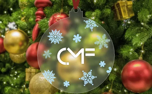 Bombka choinkowa z logo CMF, ozdobiona motywami śnieżynek, zawieszona na choince z czerwonymi i złotymi ozdobami w tle.