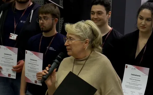 Kobieta mówi do mikrofonu podczas konferencji. Wokół niej stoją studenci, którzy trzymają dyplomy.