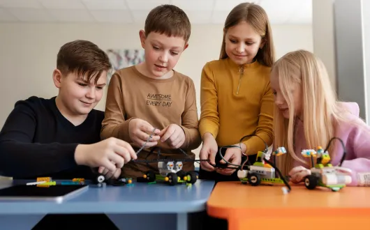 Czworo dzieci siedzi przy stoliku i wspólnie buduje modele robotów z klocków oraz przewodów.