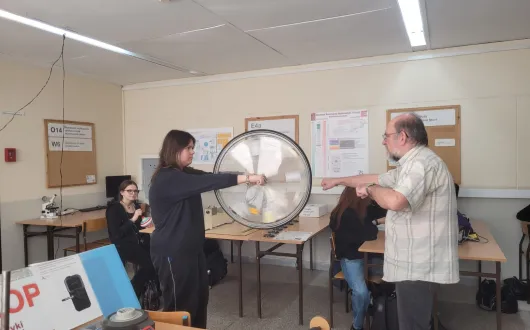 Uczennica i wykładowca stoją w laboratorium fizyki przy dużym kole do ćwiczeń z fizyki. Oboje wskazują na środek koła. W tle przy ławkach siedzą inne osoby, a na stołach leżą przyrządy pomiarowe.