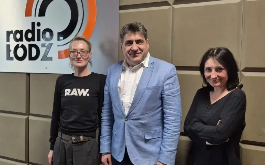 Trzy osoby stoją w studiu radiowym przy ścianie z logo Radia Łódź. W środku stoi mężczyzna w jasnoniebieskiej marynarce, a po jego bokach stoją dwie kobiety w ciemnych bluzach.