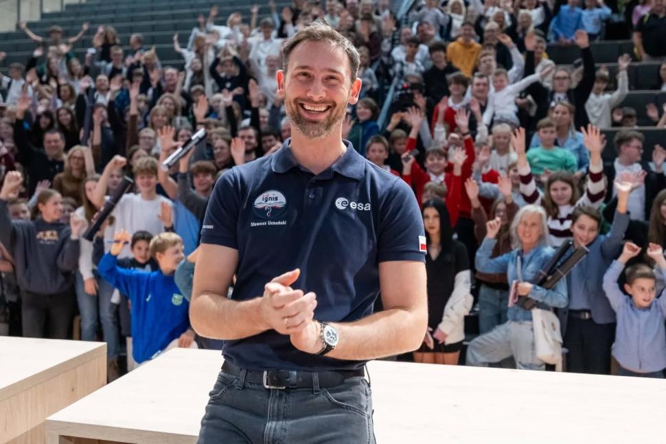 Uśmiechnięty mężczyzna w granatowej koszulce z logo ESA stoi na scenie przed dużą grupą ludzi w sali audytoryjnej. W tle widać wielu młodych uczestników, którzy podnoszą ręce i klaszczą.