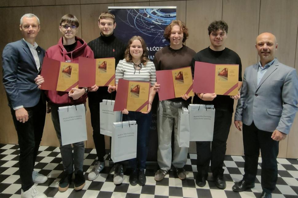 Grupa siedmiu osób pozuje do wspólnego zdjęcia na tle plakatu z logo Politechniki Łódzkiej. Pięć osób w środku, trzy kobiety i dwóch mężczyzn, trzyma dyplomy i torby z napisem „Wydział Mechaniczny Politechniki Łódzkiej”. Po bokach stoją dwaj mężczyźni w garniturach.