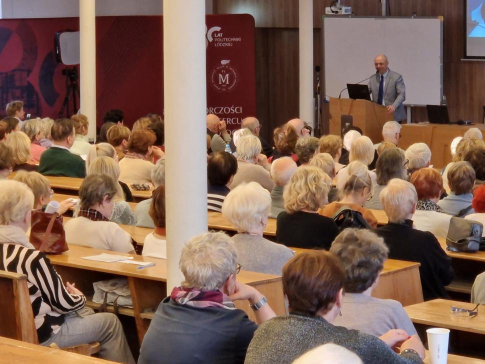 W sali wykładowej trwa wykład dla seniorów. Widzimy wiele osób siedzących na ławkach i słuchających prelegenta, który stoi przy mównicy z mikrofonem i laptopem. Za nim znajduje się tablica i ekran.