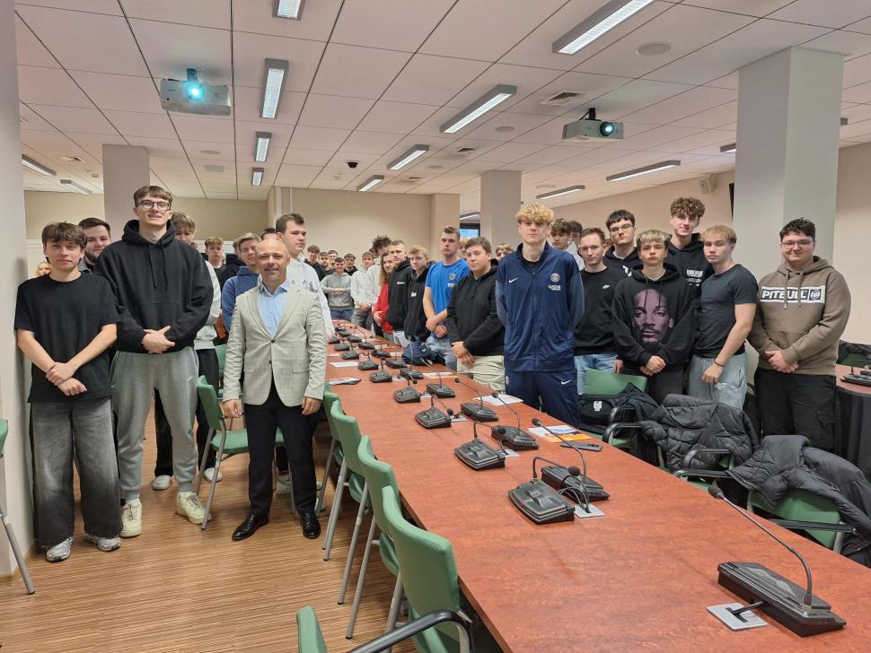 Grupa kilkudziesięciu uczniów stoi w sali konferencyjnej przy długim stole z mikrofonami. Na pierwszym planie, pośrodku, stoi mężczyzna w jasnej marynarce i koszuli. 