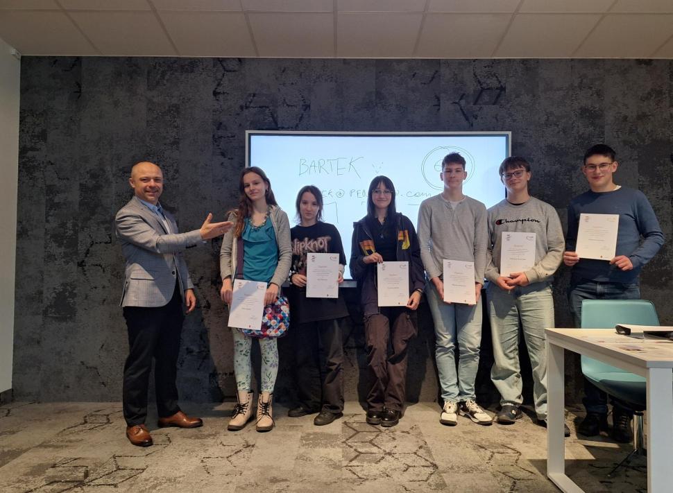 Zdjęcie grupowe w sali wykładowej przedstawiające kilka osób stojących w rzędzie i trzymających certyfikaty. Obok nich stoi prowadzący, który gestem wskazuje na uczestników. W tle widoczna jest ekran.