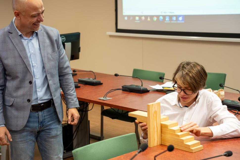 W sali wykładowej prowadzący zajęcia obserwuje ucznia podczas układania drewnianej konstrukcji schodkowej na stole. W tle widoczne są stoły konferencyjne, mikrofony i ekran projekcyjny.