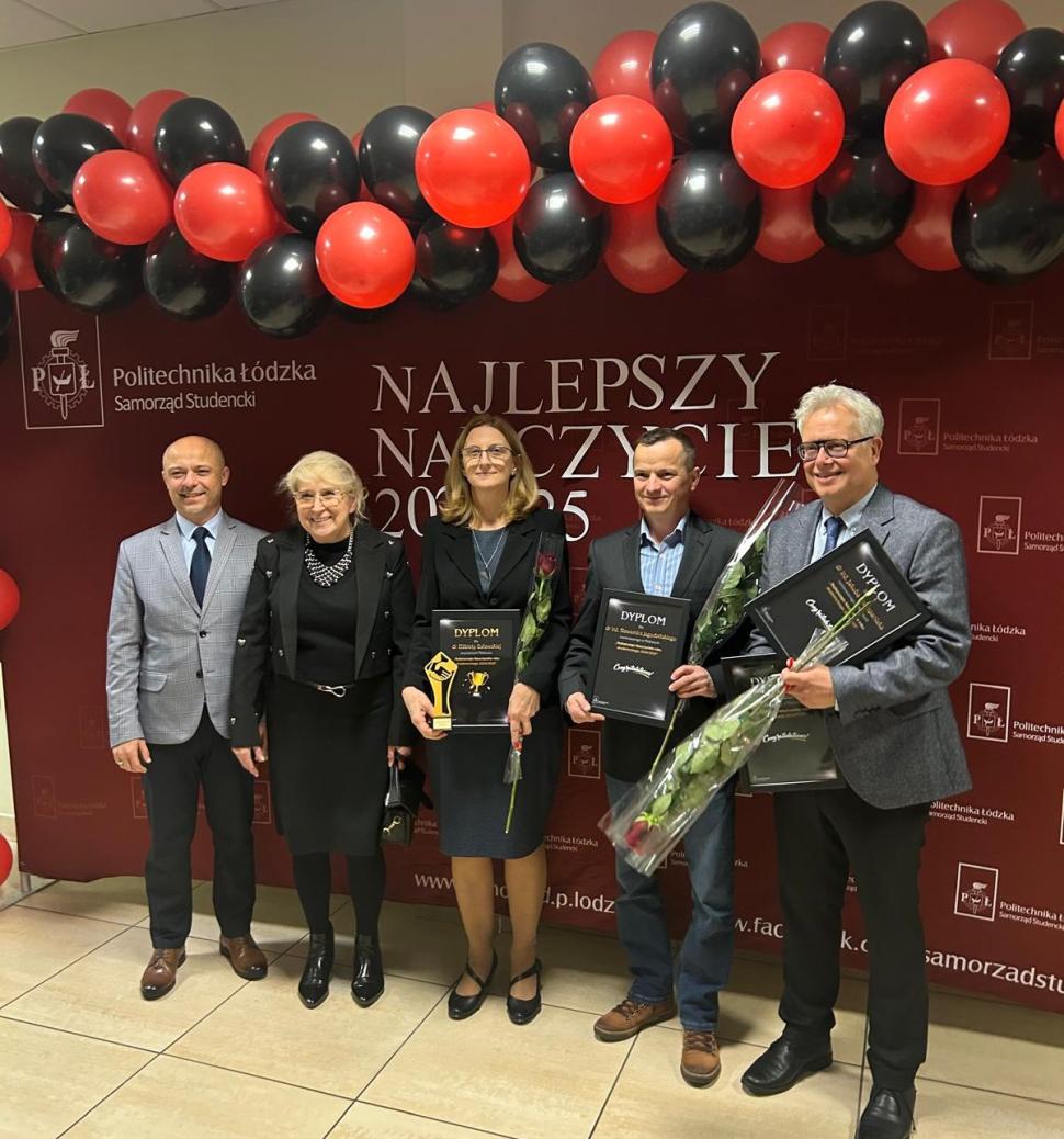 Grupowe zdjęcie kilku osób stojących przed ścianką z napisem „Najlepszy Nauczyciel”. Część osób trzyma dyplomy i kwiaty. Nad nimi widoczna jest dekoracja z czerwono-czarnych balonów.