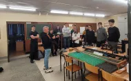 Wykładowca stoi w laboratorium fizycznym i omawia doświadczenie z urządzeniem pomiarowym. Wokół niego stoi grupa uczniów. Na stołach widać elementy zestawu do ćwiczeń i pomiarów.