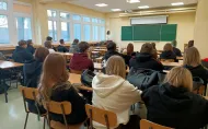 Uczniowie siedzą w sali lekcyjnej i patrzą w stronę tablicy. Przy tablicy stoi nauczycielka, która prowadzi lekcję. Na ławkach leżą plecaki i zeszyty.