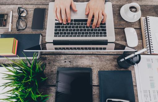 Widok z góry na biurko z laptopem, przy którym ktoś pisze na klawiaturze. Wokół znajdują się notatniki, telefon, tablet, słuchawki, kubek kawy, roślina i akcesoria biurowe.